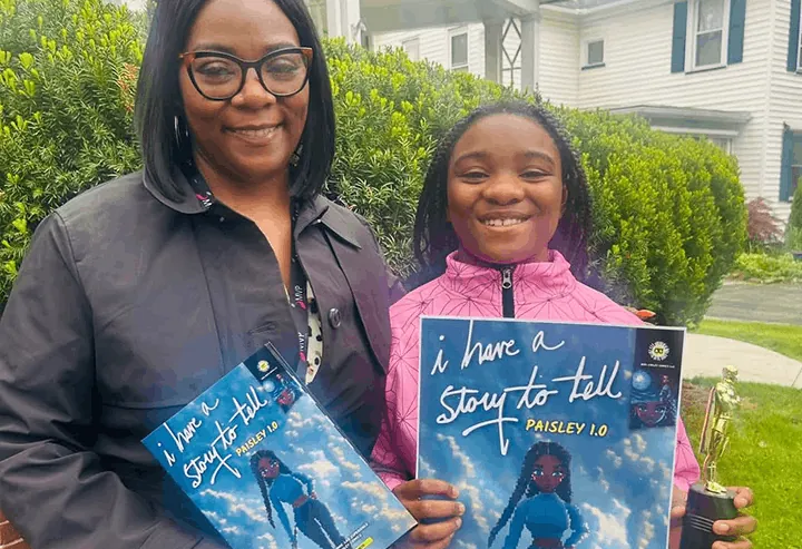 A mother and daughter outside, each holding a copy of the daughter's printed comic book, with a trophy